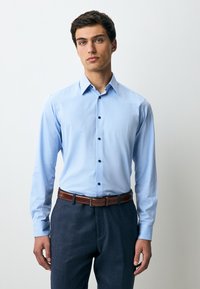 Camicia azzurro chiaro a maniche lunghe con un motivo a scacchi sottili, collo con bottoni e bottoni blu navy. Abbinata a pantaloni scuri e cintura marrone.
