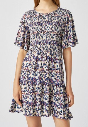 Femme portant une robe courte fleurie avec corsage smocké et manches flottantes aux couleurs bleu, violet et blanc.