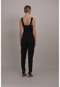 Mono negro con tirantes anchos y diseño de espalda en V. Presenta un cuerpo ajustado, piernas entalladas y zapatos de tacón alto en negro.