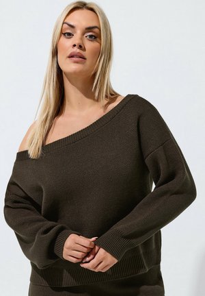 Blondynka o prostych włosach, ubrana w ciemnobrązowy, odsłaniający ramiona sweter dziany na jednolitym jasnym tle.