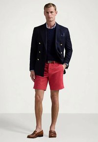 Άνδρας που φοράει ναυτικό μπλε διπλό μπλέιζερ, ναυτικό πουλόβερ, μπλε ριγέ πουκάμισο, κόκκινες βερμούδες, καφέ ζώνη και καφέ loafers, στέκεται μπροστά σε άσπρο φόντο.