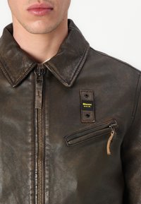 Veste en cuir marron avec un col montant, finition texturée, fermeture éclair à l'avant, patch sur la poitrine gauche avec logo, et poche latérale zippée avec tirette en cuir.