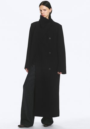 Femme aux cheveux foncés portant un long manteau noir à col montant et un pantalon noir à jambes larges, debout sur un fond blanc uni.