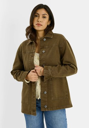 Joven mujer vistiendo una chaqueta verde oliva abotonada sobre un suéter de punto color crema y vaqueros azules, de pie contra un fondo liso y claro.