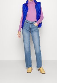Blue quilted vest over a pink turtleneck, paired with light blue jeans and yellow loafers. The outfit features a modern, casual style.