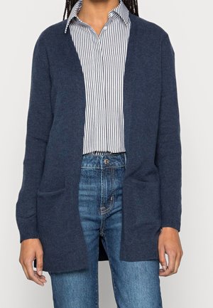Person trägt eine marineblaue offene Strickjacke über einem weiß-schwarz gestreiften Hemd mit blauen Jeans.