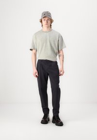 Ljusgrön, kortärmad, rundhalsad t-shirt; mörka byxor; grå keps med färgglada detaljer; svarta snörda stövlar; enspårig bakgrund.