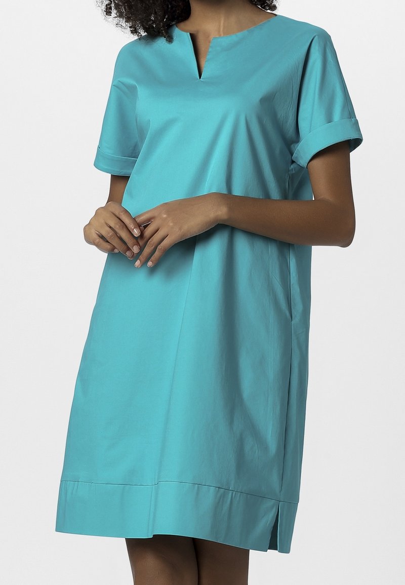 Robes en coton turquoise avec un décolleté en V, des manches courtes et des poignets roulés. Le design présente une légère forme évasée et des fentes latérales.