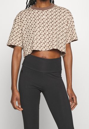 Osoba ubrana w beżowo-brązowy krótki top Adidas z geometrycznym wzorem oraz wysokowysokie czarne legginsy, stojąca na tle jednolitego tła.