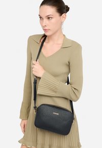 Černá krosbody taška z umělé kůže s texturovaným povrchem, zlatými doplňky a zipovým uzávěrem, nošená přes žebrované olivově zelené šaty.