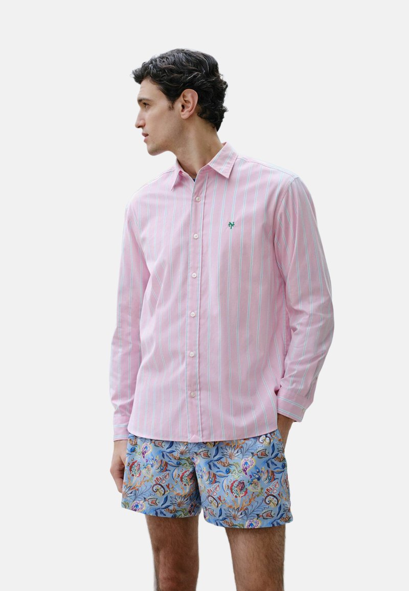 Homme aux cheveux foncés portant une chemise rose à rayures boutonnée et un short bleu à motif floral, regardant vers la gauche avec une main dans la poche.