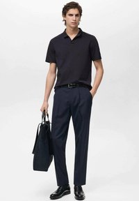 Marinblå pikétröja, mörka skräddarsydda byxor, svart bälte och blanka svarta skor, bärande en svart tote-väska. Enkel, strukturerad outfit.