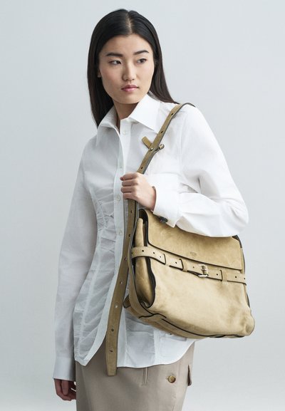 Νεαρή γυναίκα φορώντας ένα λευκό φανελάκι με ρυτίδες και μπεζ φούστα, κρατά μια μεγάλη μπεζ σουέτ τσάντα ώμου με μαύρες λεπτομέρειες και κούμπωμα.