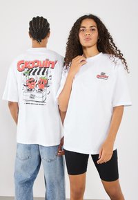 Hvid oversized t-shirt med et farverigt grafisk motiv på ryggen, der viser to tegneseriefrugter og teksten "Groovin'".