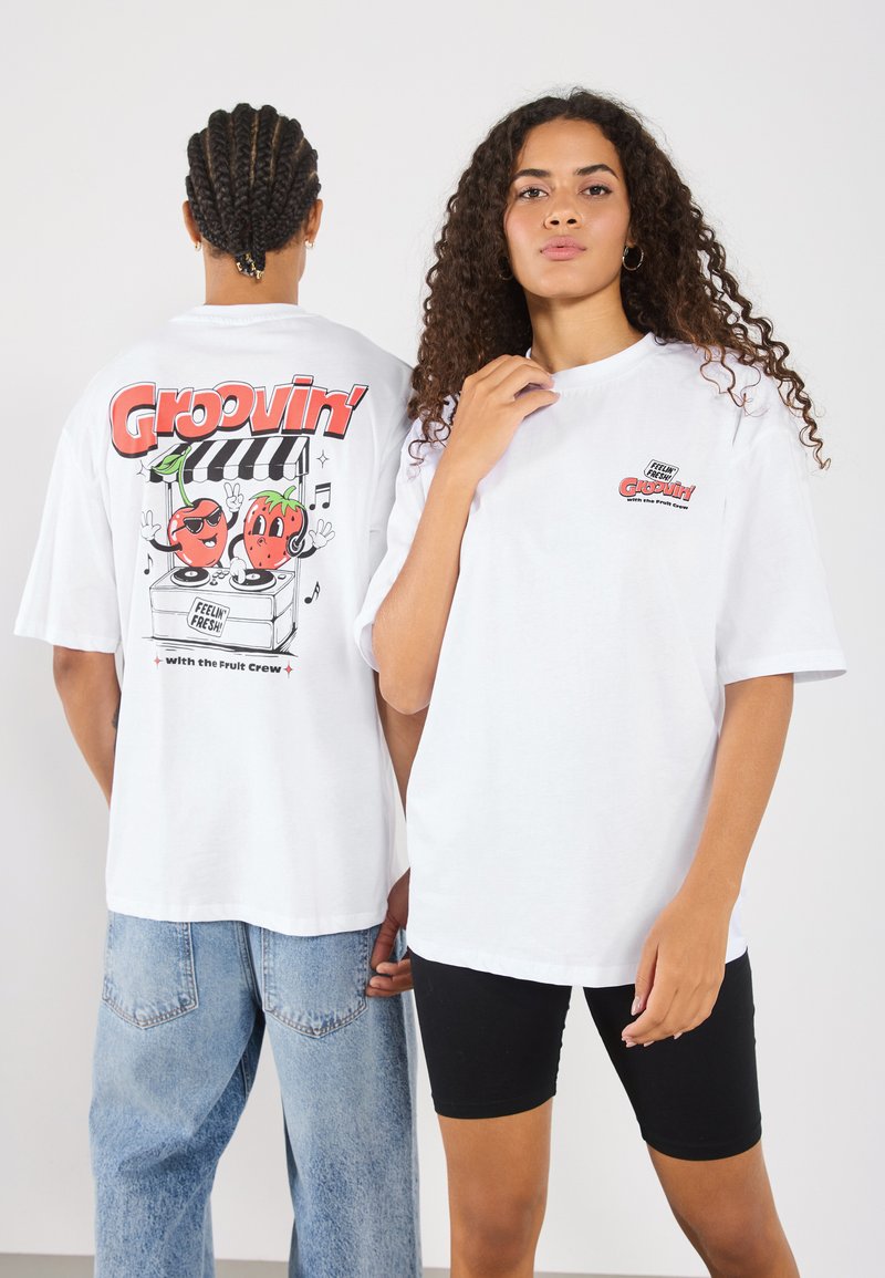 Balta per didelė t-shirts su spalvinga grafika nugaros pusėje, vaizduojančia du animacinius vaisių personažus ir tekstu "Groovin'".