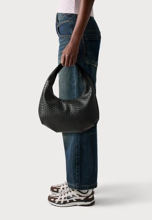 Sort vævet læder hobo taske med en afrundet form, bredt håndtag og struktureret overflade, holdt af en person iført mørke jeans og sneakers.