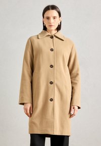Manteau en laine beige avec un col, fermeture à boutons sur le devant, poches latérales et manches longues, mettant en avant des boutons foncés comme accents.
