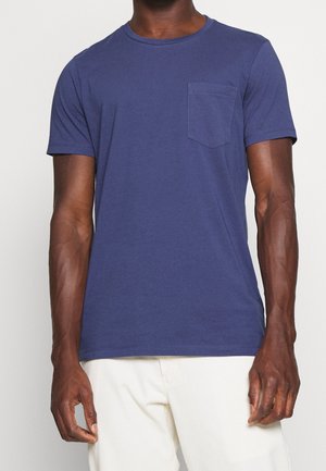 Mann trägt ein schlichtes dunkelblaues kurzärmeliges T-Shirt mit Brusttasche und cremeweißen Hosen, steht vor einem schlichten Hintergrund.