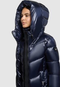 Chaqueta acolchada azul marino con textura brillante y suave, capucha alta y cierre de cremallera. Presenta un parche de logo en el hombro izquierdo.