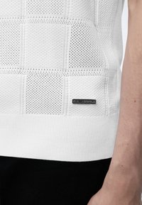 Gros plan d'un vêtement en maille blanche avec un motif carré texturé et une petite étiquette noire rectangulaire près de l'ourlet sur le côté et la main d'une personne.