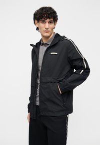 Chaqueta negra ligera con capucha, cierre de cremallera, puños elásticos y dos bolsillos laterales. Presenta franjas blancas en los lados y un pequeño parche con logo.