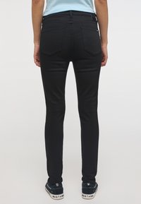 Juodieji liesieji džinsai turi lygią tekstūrą, siuvinėtas užpakalines kišenes su išlenktu dizainu ir yra derinami su tamsiais sportbačiais.