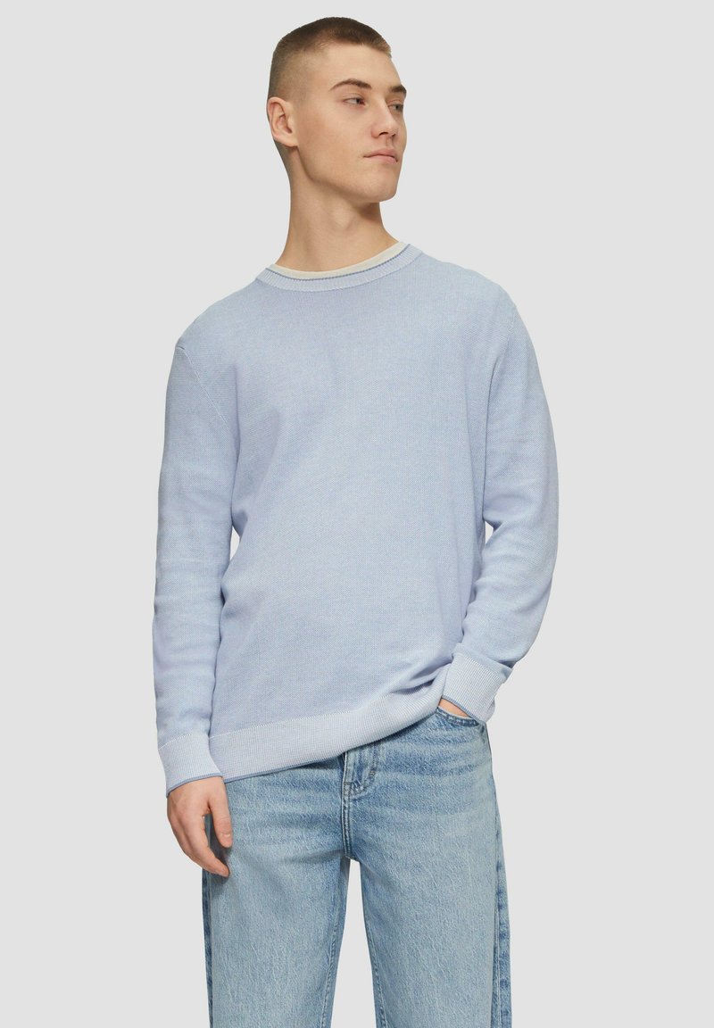 Heller blauer Strickpullover mit strukturierter Oberfläche, runder Halslinie und gerippten Manschetten. Kombiniert mit Jeans in mittlerer Waschung.