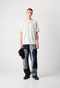 Světle zelené oversized tričko, uvolněný střih; modré džíny s díky patchworku a kontrastními odstíny; černá čepice držená v ruce; černé tenisky.