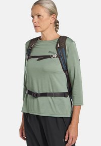 Hellgrünes Langarmhemd mit einem Logo, dazu ein braunes verstellbares Geschirr über der Brust und schwarze Gurte. Für den Außeneinsatz konzipiert.