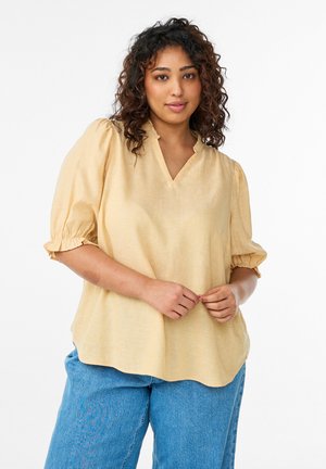 Mladá žena s kučeravými vlasmi, oblečená v svetložltej blúzke s nadýchanými rukávmi a modrých džínsoch, stojaca na bielom pozadí.