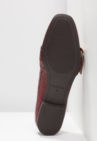 Semelle d'une chaussure mocassin texturée bordeaux avec motif antidérapant et taille 37 marquée, reposant contre une surface d'angle blanche.