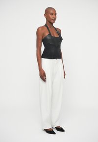 Mulher vestindo um top de pele preto sem mangas com detalhe de fivela, calças largas brancas com riscas finas, e sapatos de salto agulha pretos, de pé contra um fundo branco.