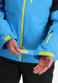 Giacca impermeabile blu con accenti gialli, tasche con zip e polsini regolabili in Velcro. Realizzata in materiale liscio e resistente.