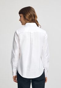Camicia bianca a maniche lunghe con colletto classico, dettaglio sulla spalla posteriore, orlo curvo e polsini con bottoni. Tessuto morbido e liscio.