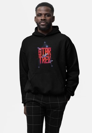 Mężczyzna ubrany w czarną bluzę z kapturem z czerwonym napisem "Star Trek" i grafiką statku kosmicznego, w połączeniu z czarnymi spodniami z białymi liniami siatki.