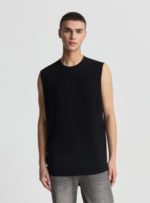 Młody mężczyzna z krótkimi włosami, ubrany w czarną koszulkę bez rękawów i szare dżinsy, na tle jednolitego jasnoszarego tła.