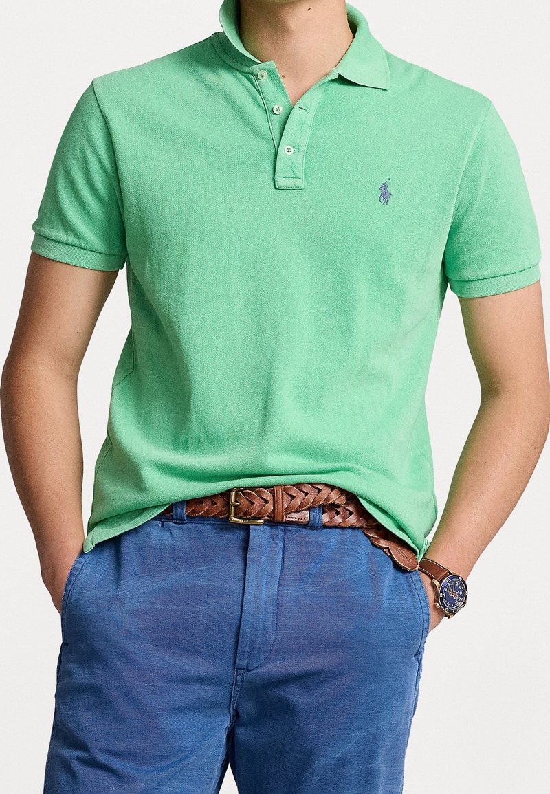 Personne portant un polo vert, un pantalon bleu, une ceinture en cuir tressé marron et une montre-bracelet avec un cadran bleu et un bracelet marron.