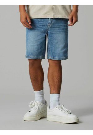 Osoba má na sebe svetlomodré riflové kraťasy, biele členkové ponožky a biele tenisky a stojí na sivej podlahe.