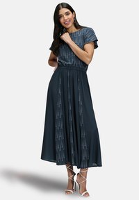 Donkerblauwe maxi-jurk met korte mouwen, elastische taille en verticale patronen. Gladde stof valt elegant tot de vloer. Witte sandalen met bandjes.