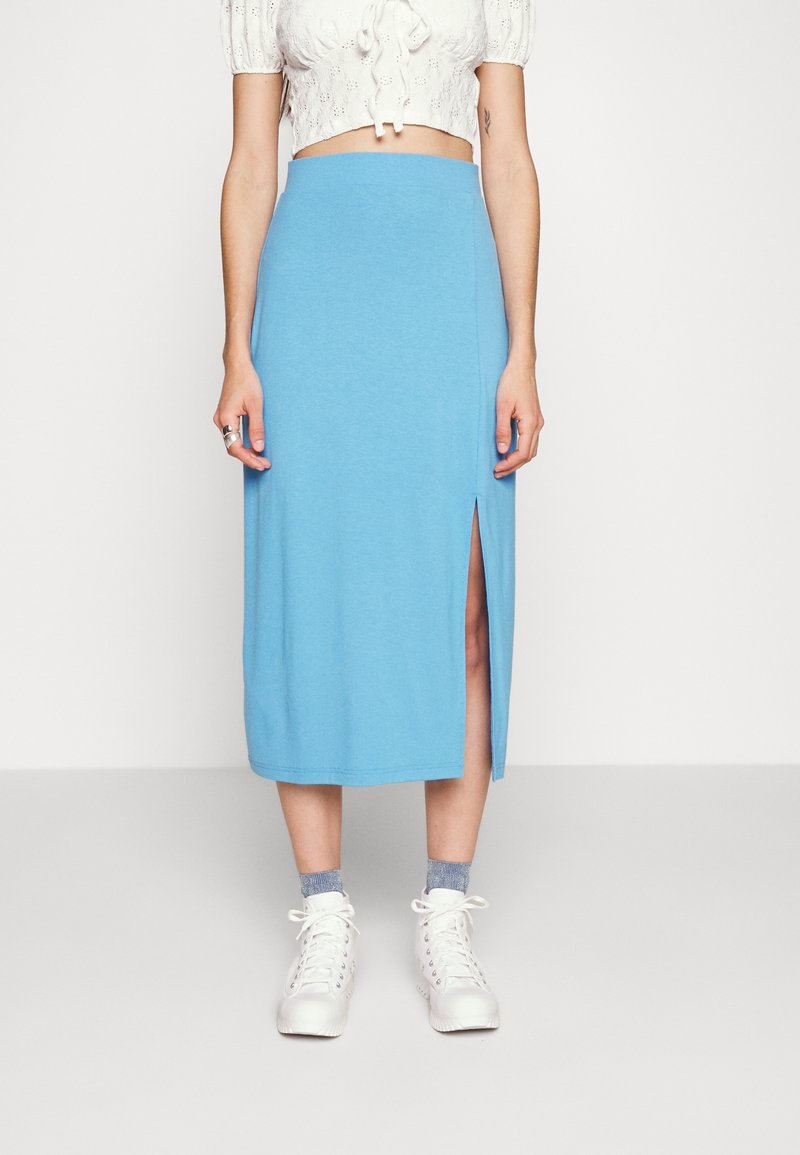 Femme portant une jupe midi bleue avec une fente latérale, un chemisier blanc court, des chaussettes grises et des bottines blanches à lacets, debout sur un fond uni.