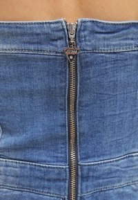 Lähikuva hopeisesta Guess-logovetoketjusta sinisessä farkkavaatteessa, jossa näkyy ompelu ja pieni vilaus paljasta ihoa ylhäällä.