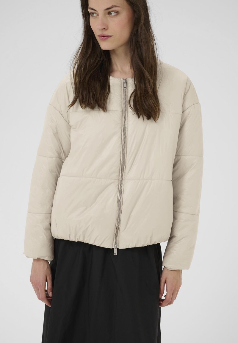Doudoune beige clair, dotée d'une fermeture éclair frontale, d'un col arrondi et d'un ourlet côtelé. Tissu lisse avec une texture légèrement brillante.
