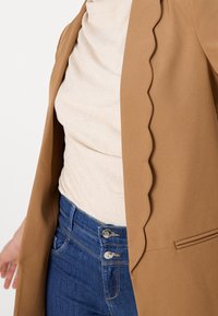 Gros plan d'une personne portant un haut beige, un blazer marron à bords festonnés et un jean bleu avec deux boutons argentés à la taille.