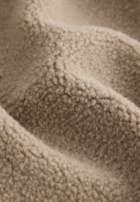 Tan fleece-tyg som visar en texturerad yta med ett mjukt, fluffigt utseende. Materialet har en subtil djup och värme.