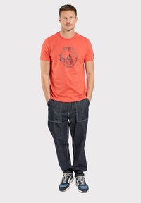 T-shirt rouge avec un motif de voilier, associé à un jean baggy bleu foncé avec des poches avant et des surpiqûres contrastantes. Des baskets complètent la tenue.