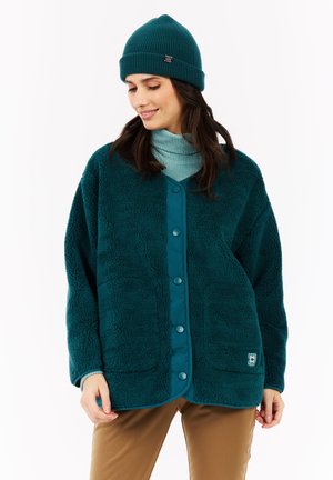 Femme portant une veste en polaire teal à boutons, un bonnet côtelé assorti, un col roulé bleu clair et un pantalon marron, regardant vers le bas avec un léger sourire.