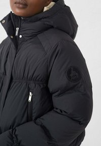 Zwarte pufferjas met capuchon, voorzien van een ritssluiting aan de voorkant, elastische manchetten en een ronde logopatch op de mouw. Gladde, geïsoleerde stof.