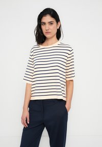 Ριγέ T-shirt σε ανοιχτή ροδακινί και σκούρο μπλε ναυτικό, με κοντά μανίκια και στρογγυλή λαιμόκοψη, σε άνετη εφαρμογή. Συνδυασμένο με σκούρες μπλε ναυτικές παντελόνες.