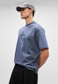 Siva kapa i plava pamučna majica s kratkim rukavima. Majica ima crni "Calvin Klein" logo na prsima. Model nosi crne hlače.