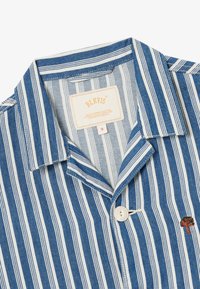Camicia a righe a maniche corte in denim blu e bianco. Presenta una chiusura con bottoni e un logo ad albero ricamato sul lato sinistro.
